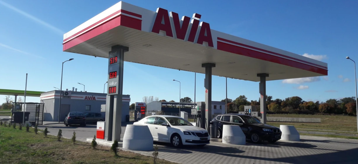 Widok z tyłu samochodu subkompaktowego na stacji benzynowej, z odbiciem lusterka bocznego i zderzaka widocznym. Pokazane są różne ceny paliwa i marka AVIA.